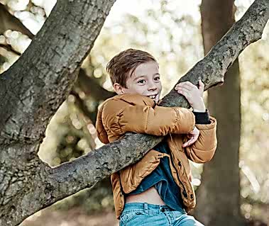 A child wearing a brown jacket and blue jeans smiles while climbing and hugging a tree branch, capturing a moment of joy. Trees and foliage are visible in the background, perfect for any family photo album. This candid scene could have been effortlessly captured by a skilled photographer in Thanet.
