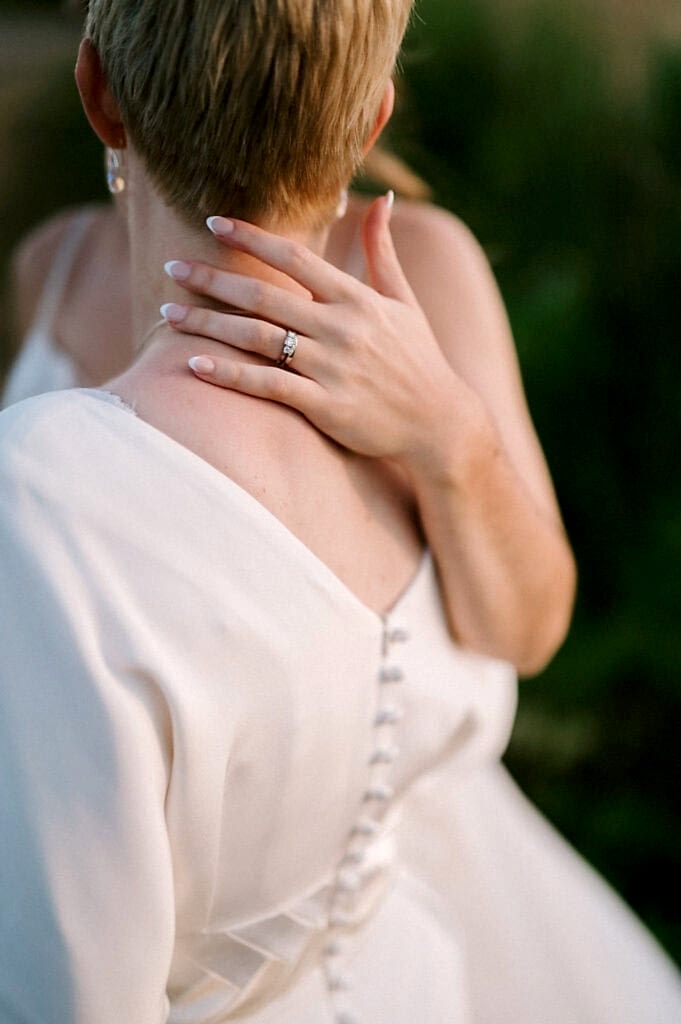 A person with short, blonde hair is seen from behind, wearing a white dress with buttons along the back. Their left hand, adorned with a ring, is gently placed on their neck. The background is blurred greenery. Image by Pearce Wedding Photography.