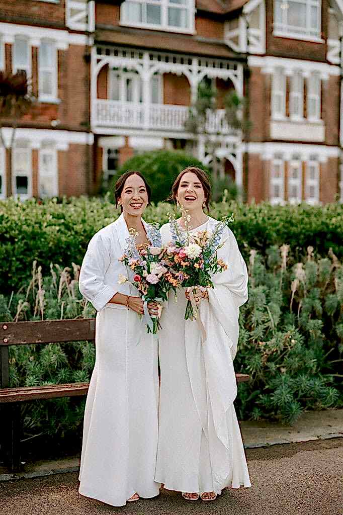 Two women in white dresses stand together outdoors, holding colorful flower bouquets. They are smiling, framed by the lush greenery and the historic charm of Ramsgate Registry Office as their backdrop. Image by Pearce Wedding Photography.