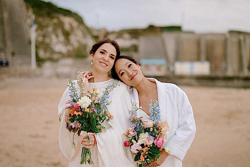 Two brides smiling with bouquets on beach.