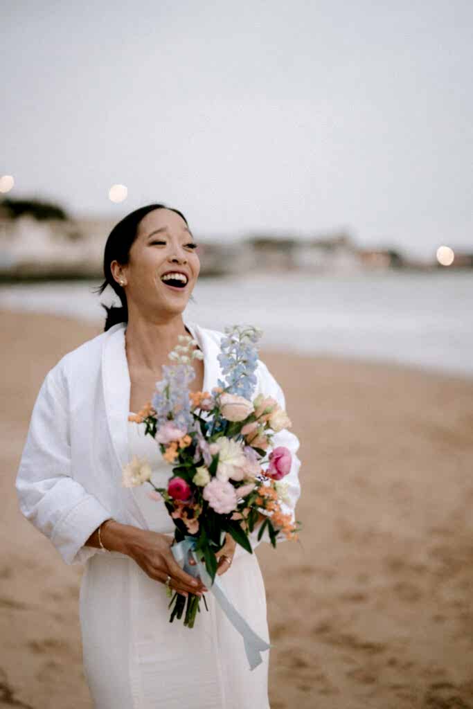 A joyful bride in a white dress and robe laughs while holding a colorful bouquet during her Ramsgate wedding. She stands on a sandy beach with gentle waves and distant buildings in the background. Image by Pearce Wedding Photography.