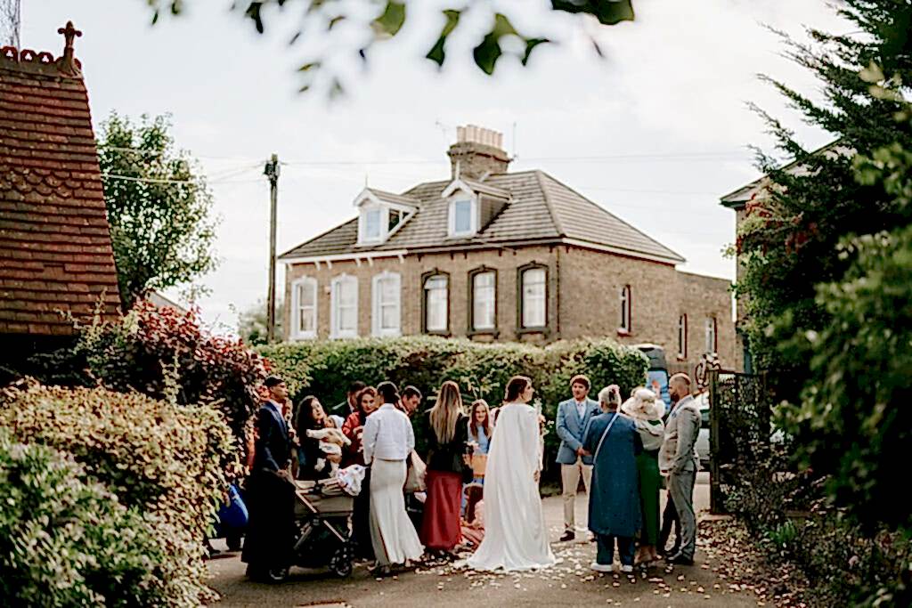 Outside the historic Ramsgate Registry Office, a group of people dressed in formal attire gather near a large brick house. Some are holding babies, and there is a stroller among them. The scene is festive, with petals scattered on the ground and trees lining the path. Image by Pearce Wedding Photography.