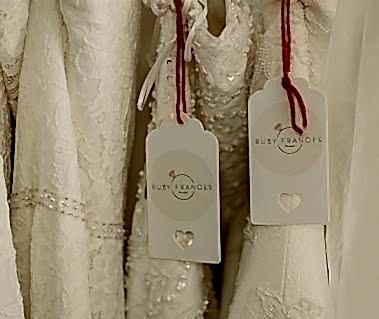 A close-up of two white lace garments with red stringed tags labeled "Ruby Frances" hanging side by side in a quaint Thanet bridal shop.