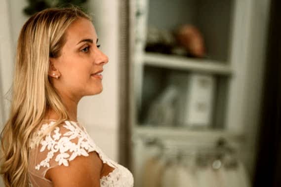 Side profile of a woman with long blonde hair, wearing a white lace dress, standing indoors near a closet with hanging clothes in a bridal shop.