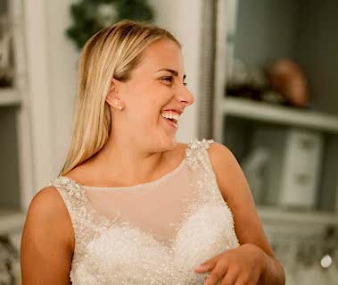 A woman with long blonde hair wearing a white sleeveless dress smiles and looks to her right. The background, reminiscent of a charming Thanet bridal shop, shows shelves with various items.