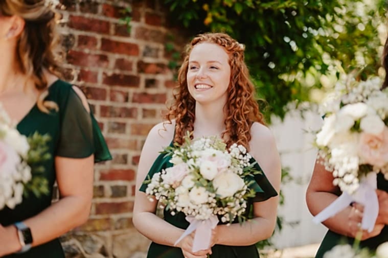 A woman with curly red hair in a dark green dress holds a bouquet of white and pink flowers, standing outdoors near a brick wall and foliage, smiling in a relaxed manner as the photographer captures the moment.
