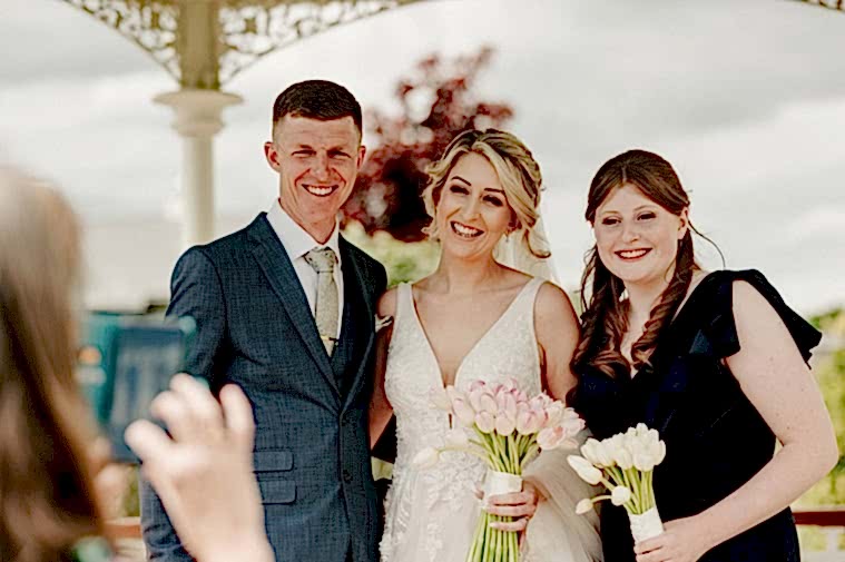Three people are posing for a relaxed photo at an outdoor event. One person is dressed in a dark suit, one in a white dress holding pink flowers, and one in a dark dress holding white flowers, capturing the essence of the wedding beautifully for the photographer.
