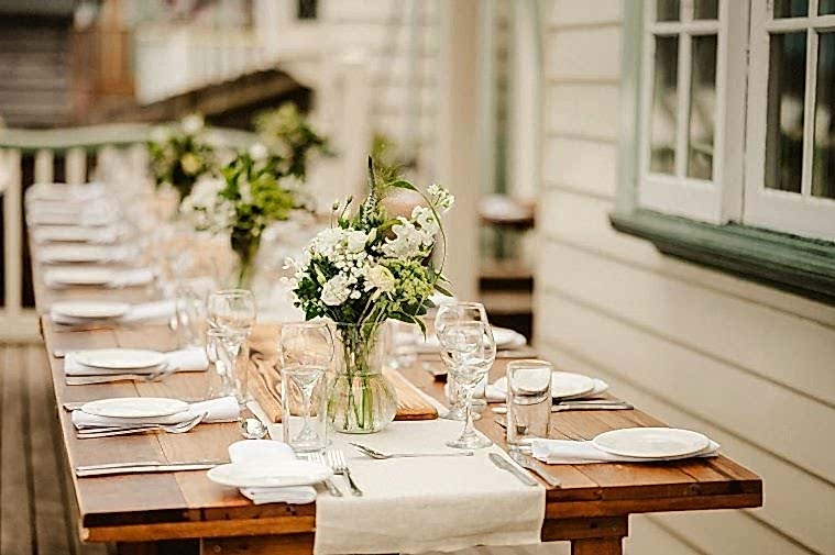 A long wooden table set for a formal meal, adorned with white plates, glassware, cutlery, light-colored napkins, and flower arrangements crafted by a local florist in glass jars, is on an outdoor patio next to a building in Kent.
