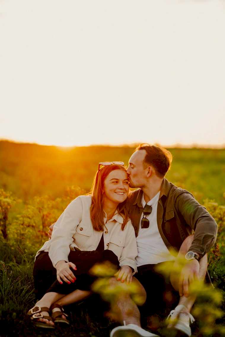 A man and woman sit together in a grassy field during sunset, with the picturesque backdrop of Botany Bay enhancing the moment. The man kisses the woman's forehead while she smiles. Both are casually dressed and wearing sunglasses, capturing their happiness as if in a serene photography session.