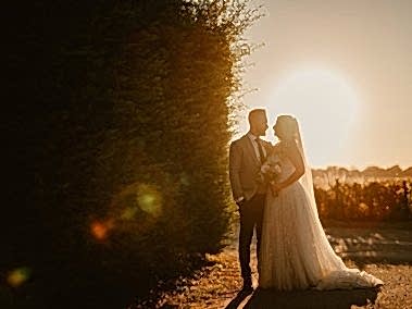 A photographer captures a bride and groom standing close together, silhouetted by the setting sun next to a tall hedge. The bride holds a bouquet and wears a flowing gown with a veil, adding to the magic of their wedding day.