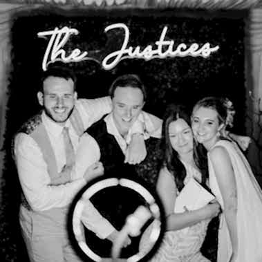 Black and white photo of four people posing in front of a sign that reads "The Justices." A ring light, often used by photographers, is visible at the bottom of the image.
