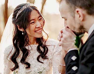 A bride smiles as a groom kisses her hand during an outdoor wedding ceremony. She wears a white dress and veil, while he wears a black suit, perfectly captured by the wedding photographer.