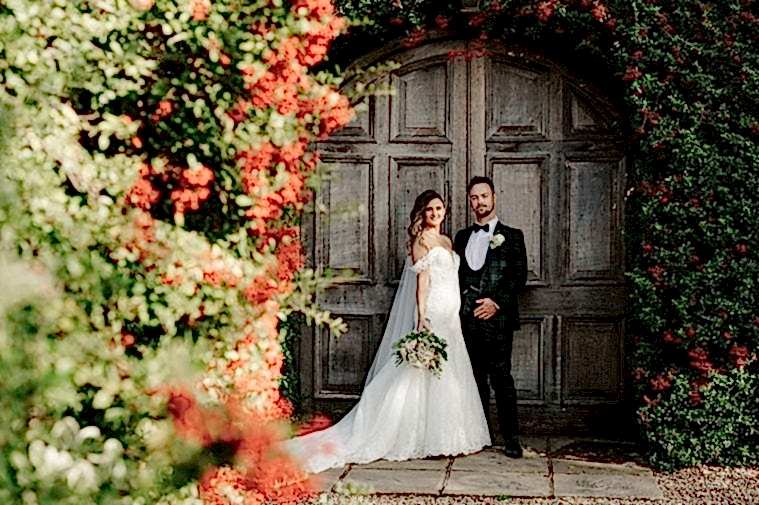 A bride and groom stand in front of a large, wooden door surrounded by red flowers and greenery. The bride holds a bouquet and wears a white wedding dress; the groom wears a black tuxedo as the wedding photographer captures their special moment.