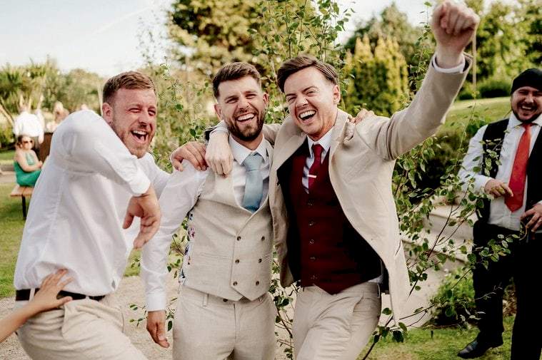 Three men in suits, two in beige and one in white, laugh and embrace joyfully outdoors. Other people are visible in the background, and trees and greenery surround them—captured perfectly by a wedding photographer.