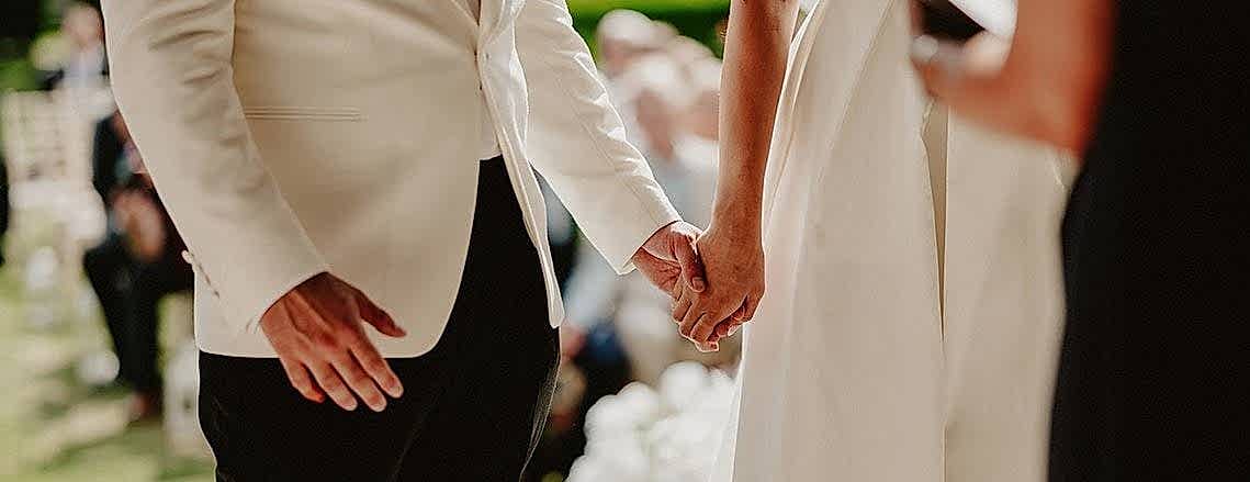 A couple holds hands at an outdoor wedding ceremony, both dressed in formal white attire, as the wedding photographer captures the moment.