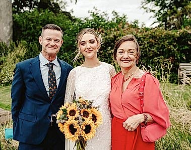 Three people are posing for a photo outside, likely taken by a wedding photographer. The person in the center, wearing a white dress and holding a bouquet of sunflowers, stands between two others: one in a blue suit on the left and another in a red outfit on the right.