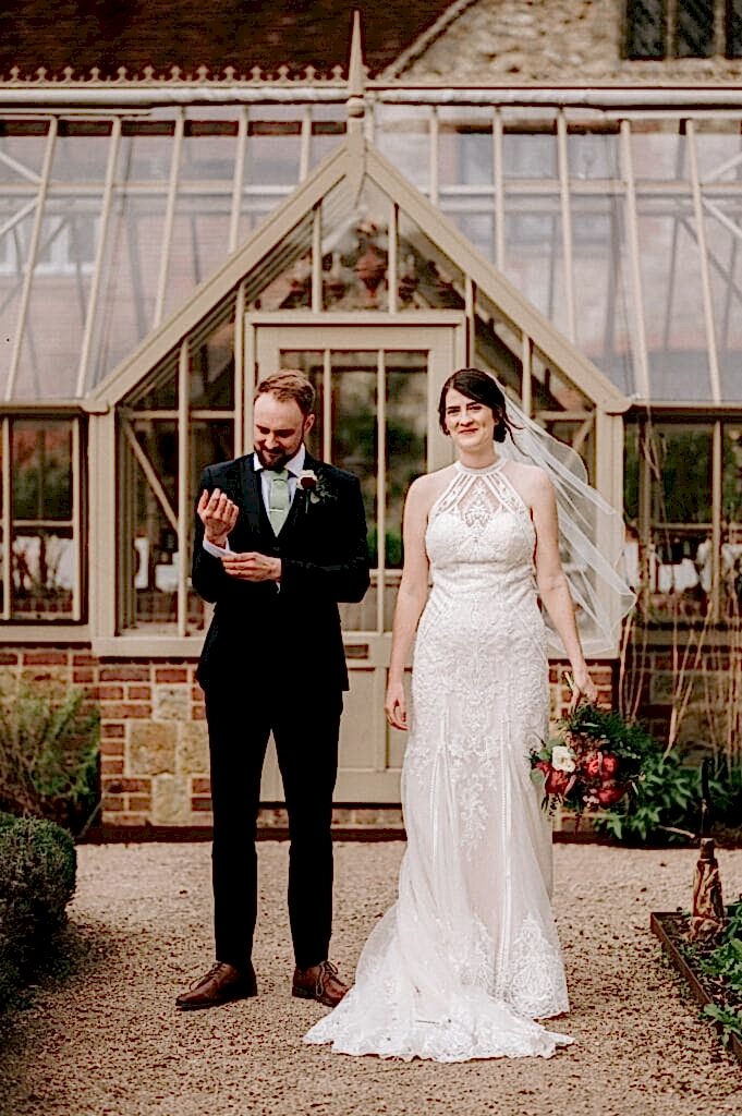 A bride in a lace gown holds a bouquet while standing next to a groom in a black suit adjusting his sleeve. They are in front of the glass greenhouse at a charming Kent wedding venue.