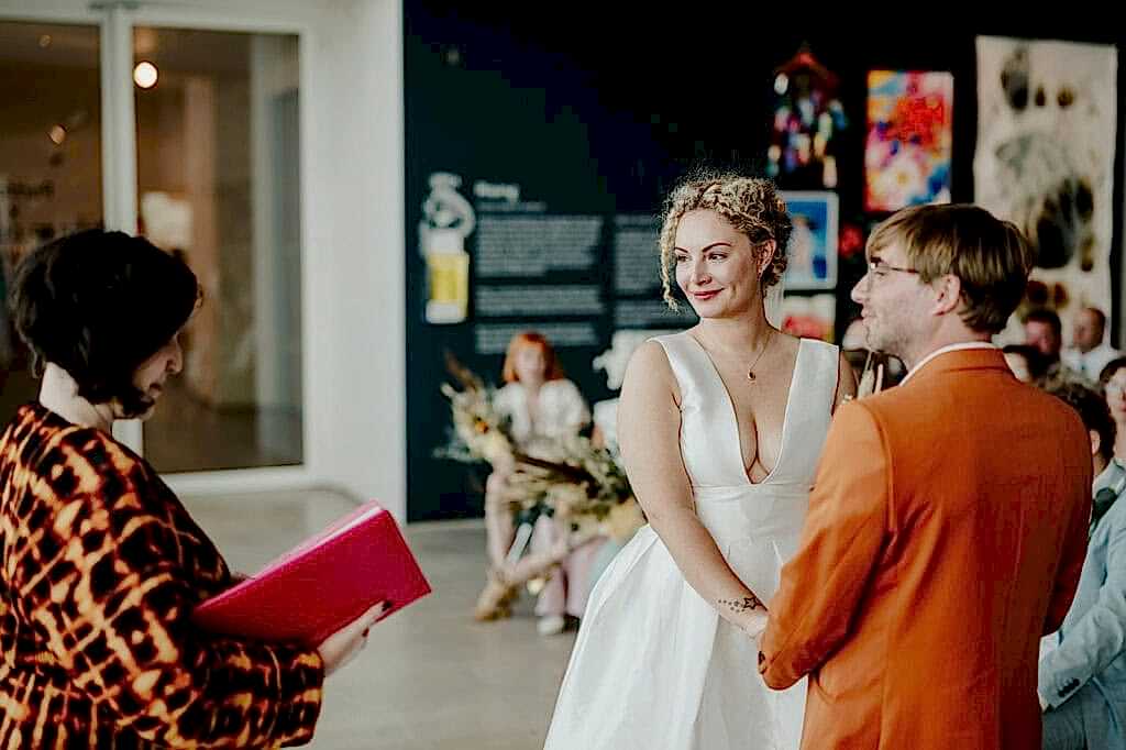 A couple stands facing each other, holding hands, during their wedding ceremony. A person officiates the ceremony, holding a pink folder. Colorful artwork is displayed on the walls in the background.