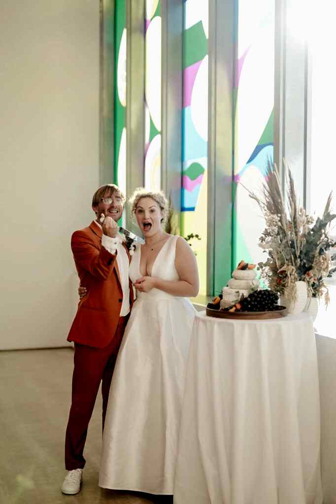 A couple stands by a table with a wedding cake. The woman in a white dress and the man in an orange suit are smiling and looking at the camera, holding their left hands up with the rings visible.