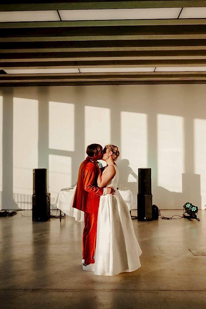 Two people, one in a red suit and the other in a white dress, share a kiss on an empty dance floor with large windows in the background, casting long shadows from the evening sun.