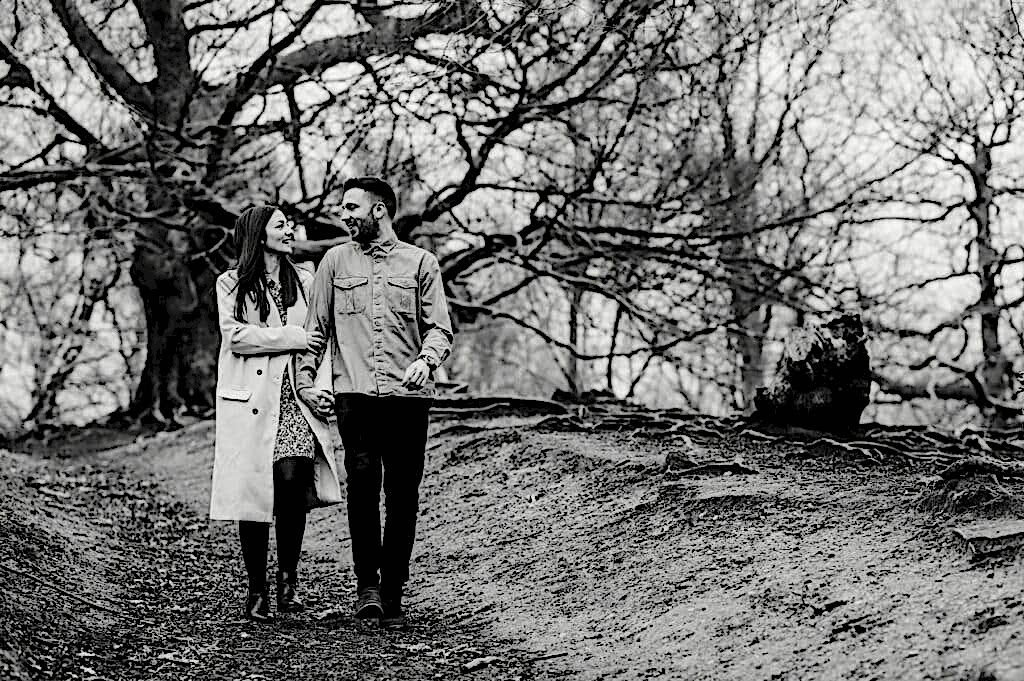 A couple walks hand in hand on a forest path lined with trees. The woman wears a long coat and the man wears a jacket and dark pants. They look at each other affectionately. The scene is in black and white.