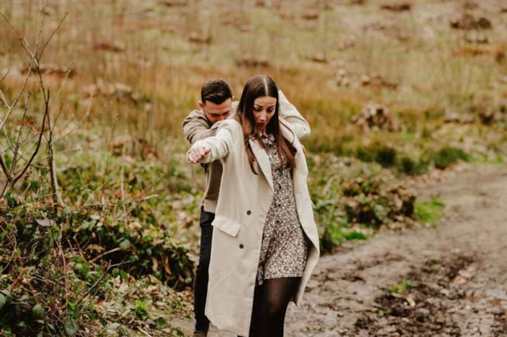 A man and a woman walk down a muddy path in a grassy field. The woman wears a beige coat and floral dress, while the man, slightly behind her, appears to guide her by holding her shoulders.