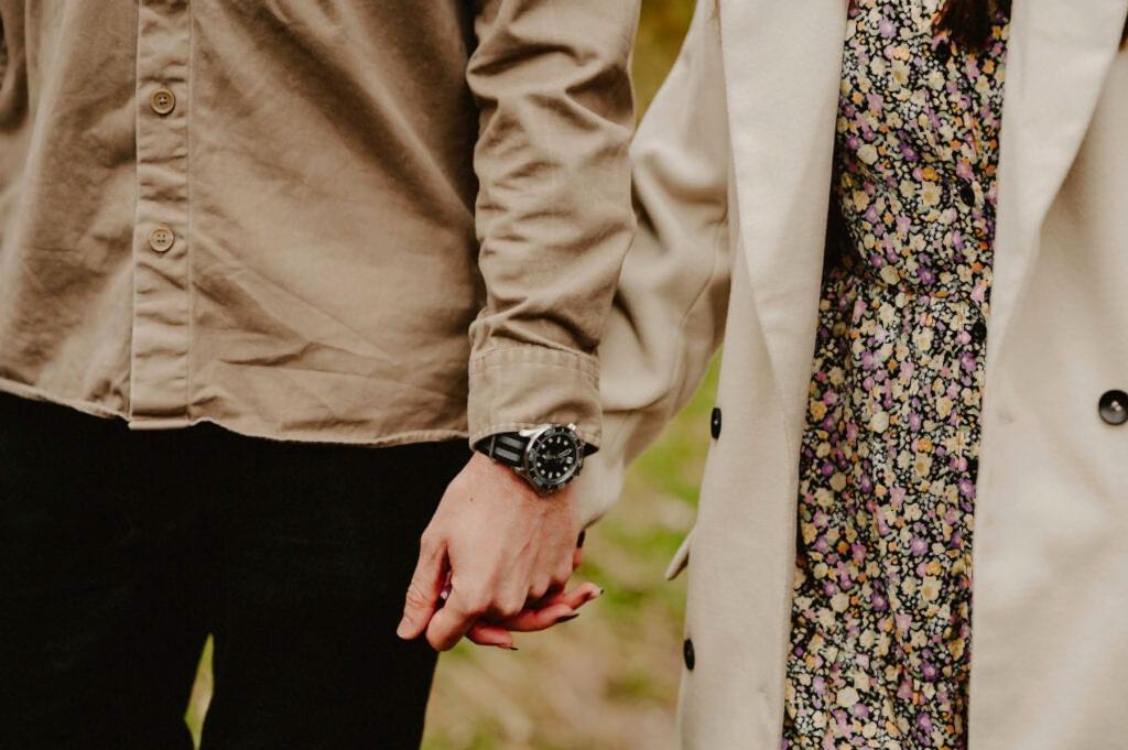 Two people are holding hands. One is wearing a beige shirt and black pants, while the other is wearing a floral dress and white coat.
