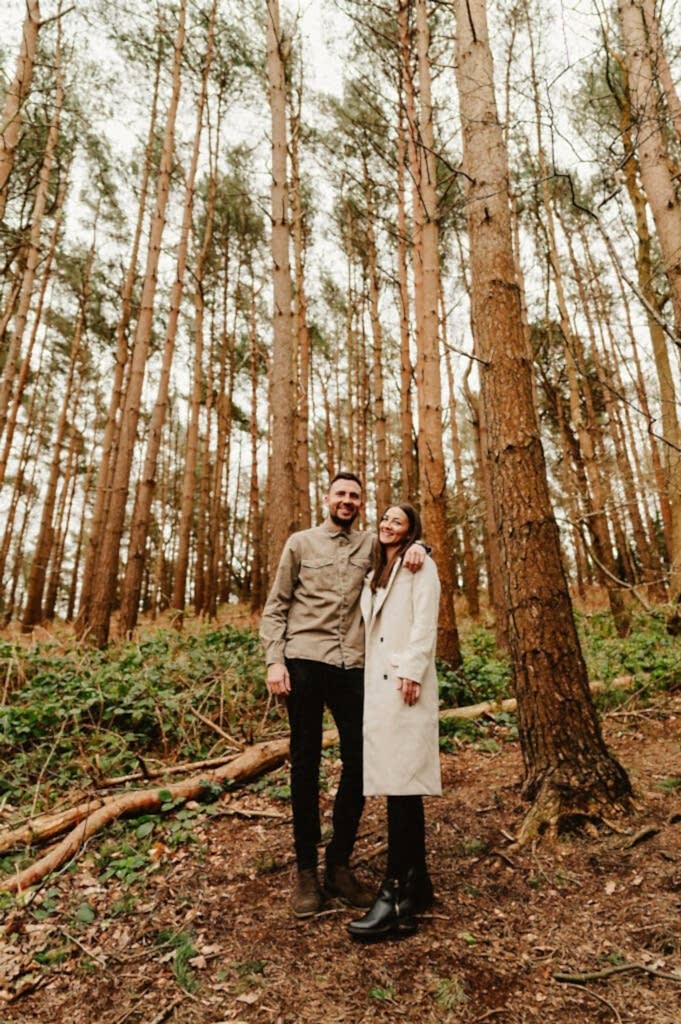 A man and a woman stand side by side in a forest. The man is wearing a khaki jacket and black pants, while the woman is in a light long coat and a beanie. Tall trees fill the background.