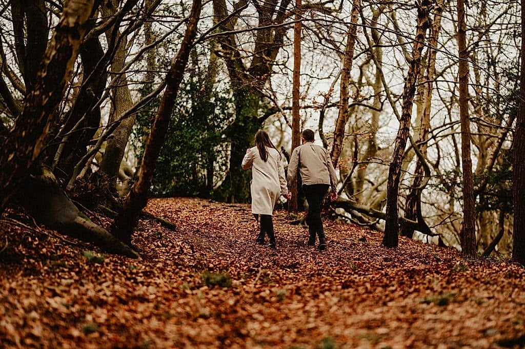 Two people holding hands walk through a forest with fallen leaves on the ground, surrounded by bare trees.