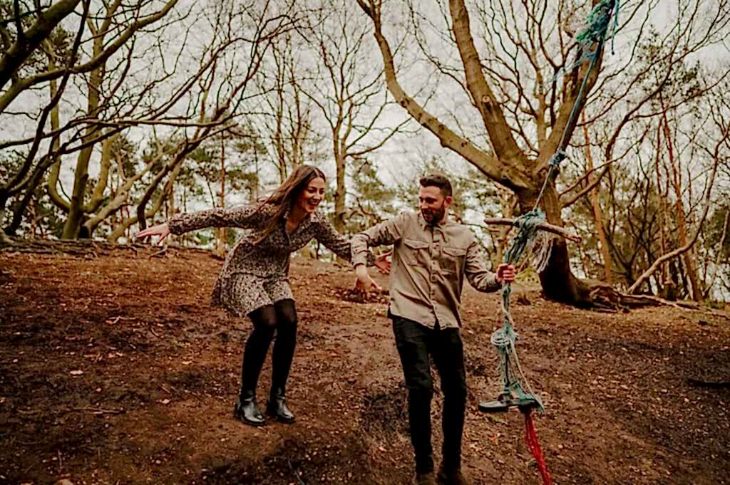 A man and woman are playing on a makeshift rope swing in a forested area, with trees and an earthy ground visible. The woman appears to be jumping or balancing while the man steadies the swing.