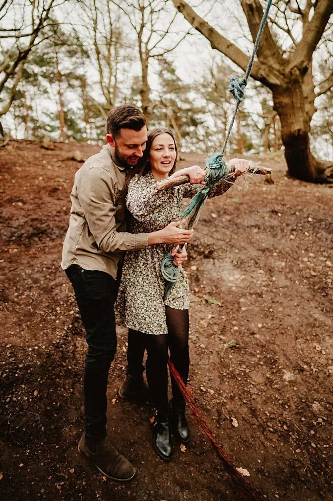 A man and woman in a wooded area, standing on a dirt slope, holding onto a thick rope tied to a tree branch above. The man is behind the woman, helping her grip the rope.