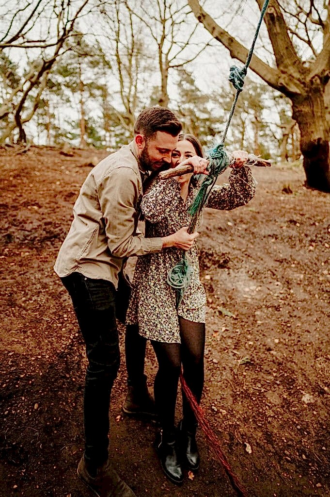 A person in a floral dress holds onto a rope swing with another person in a beige jacket standing behind them, in a wooded area.