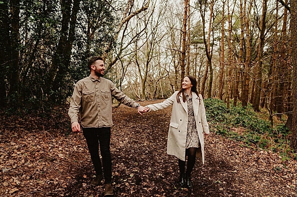 A man and woman holding hands, walking through a forest path covered in leaves, surrounded by trees and greenery.