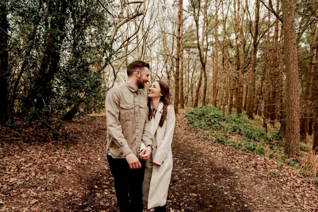 A couple walks hand in hand on a leaf-covered path through a forest, smiling at each other.