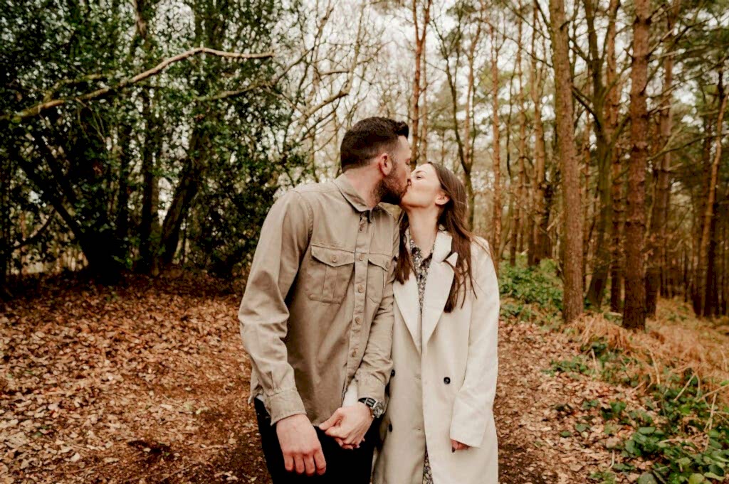 A couple, holding hands and wearing light jackets, kisses while walking on a leaf-strewn path through a dense forest.