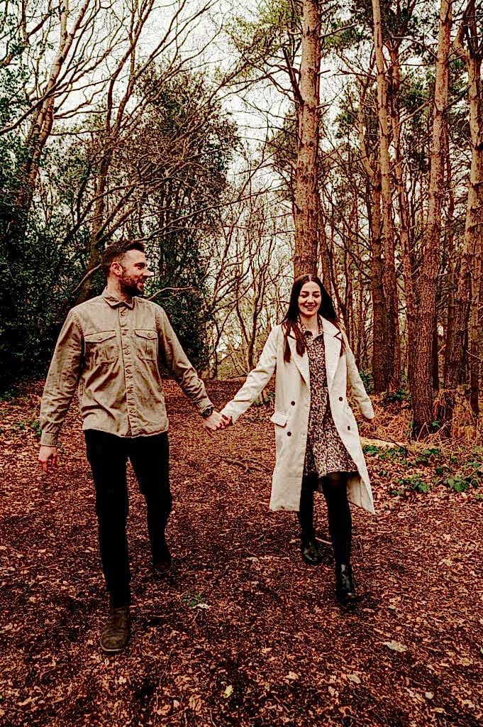 A man and woman hold hands while walking on a forest path covered with fallen leaves, surrounded by tall trees. They are both smiling and dressed in casual attire.