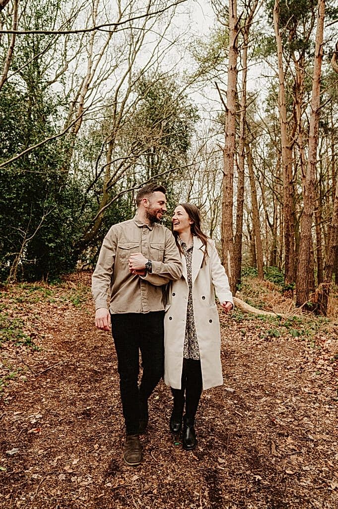 A man and woman walk hand-in-hand through a wooded area with trees and dry leaves on the ground. Both are smiling and wearing coats.