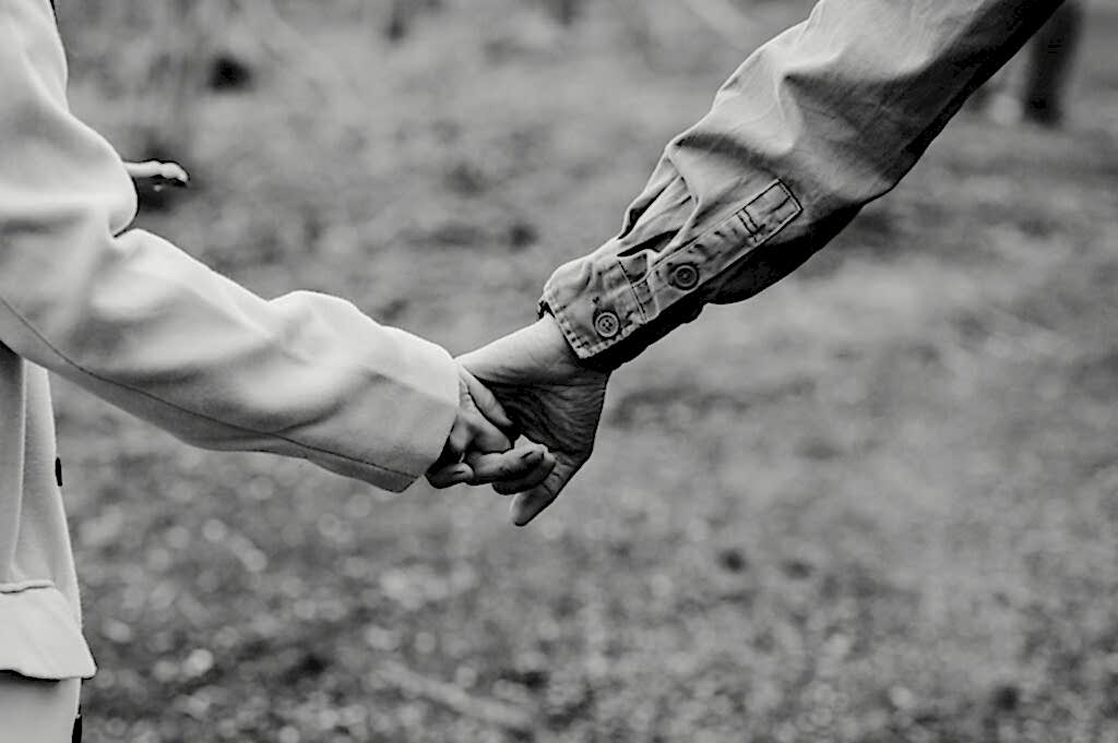 Two people holding hands outdoors, one wearing a light coat and the other in a darker jacket.