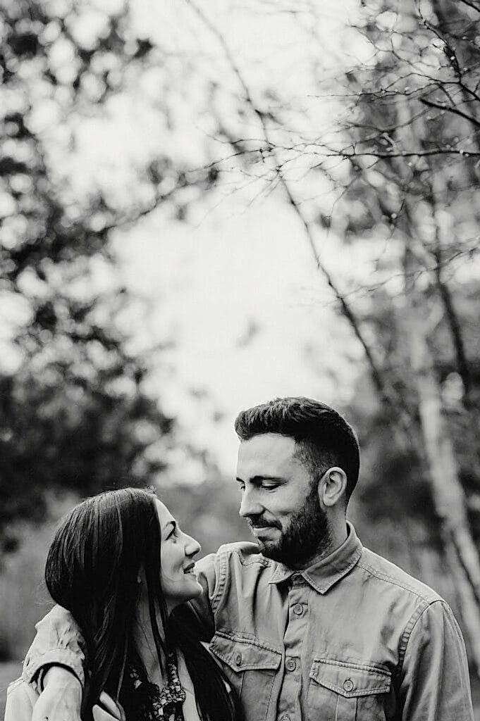A black-and-white photo of a man and woman standing closely together outdoors, looking affectionately at each other. Trees and foliage are blurred in the background.