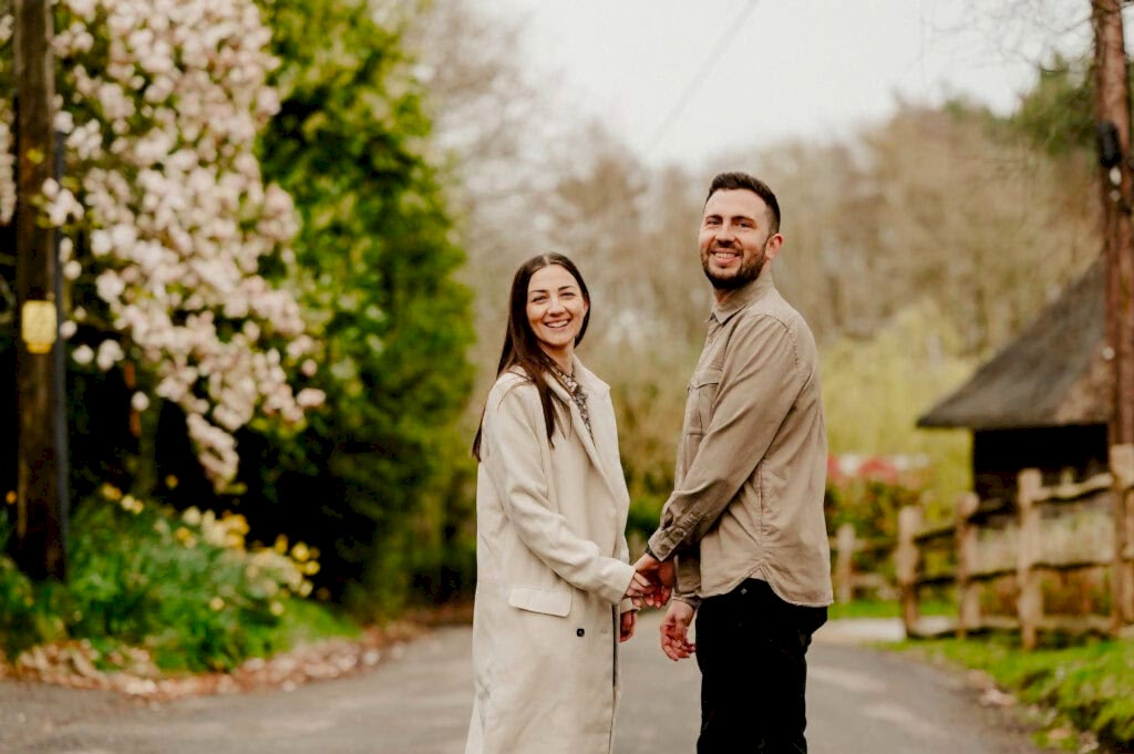 A man and woman stand holding hands, smiling at the camera on a tree-lined street with blooming flowers. The woman wears a light coat, and the man is dressed in casual attire.