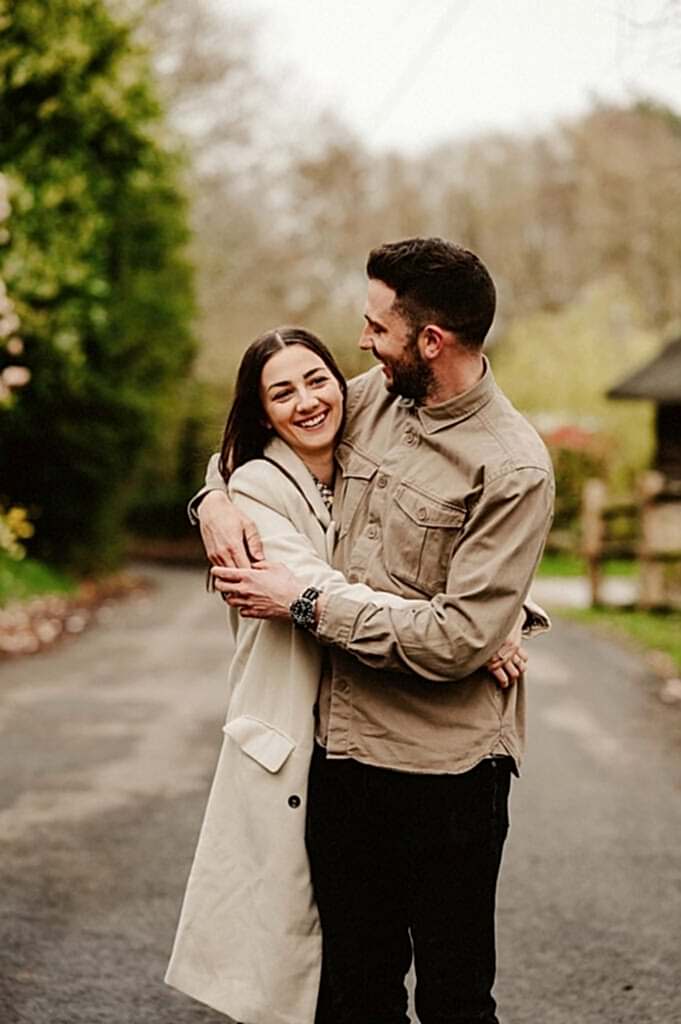 A man and a woman stand in the middle of a rural road, smiling and embracing. The background features trees and a wooden fence.