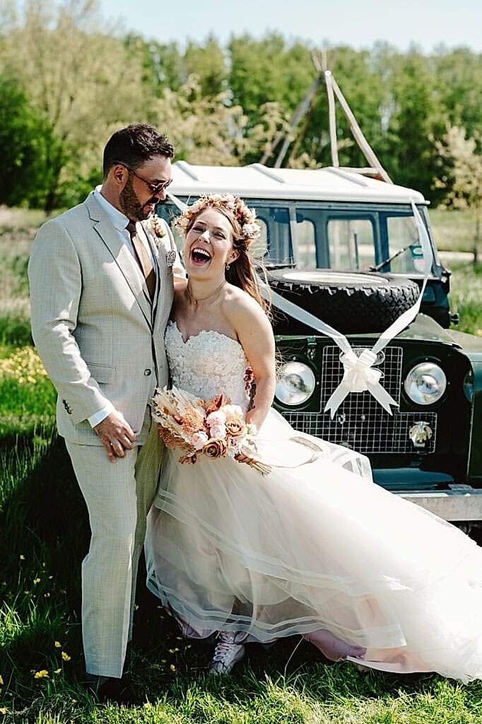 A bride and groom stand smiling and laughing in front of a decorated vintage off-road vehicle, with greenery and trees in the background. The bride holds a bouquet and wears a floral headpiece.