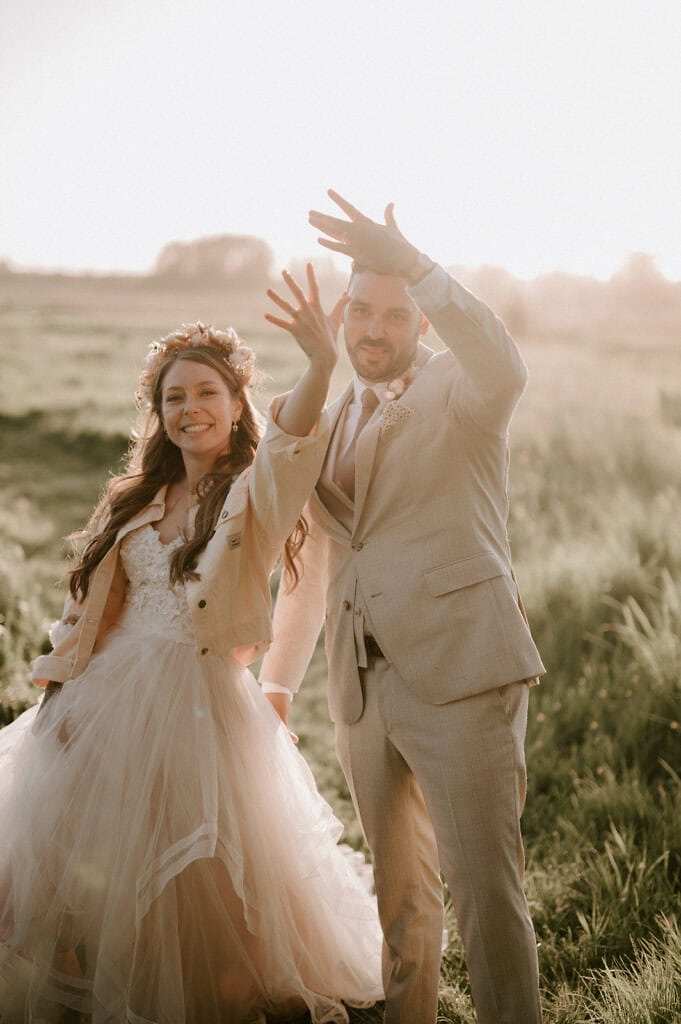 A bride and groom are standing outdoors in a grassy field, wearing wedding attire. Both are smiling and waving their hands toward the camera. The scene is lit by warm, natural light.