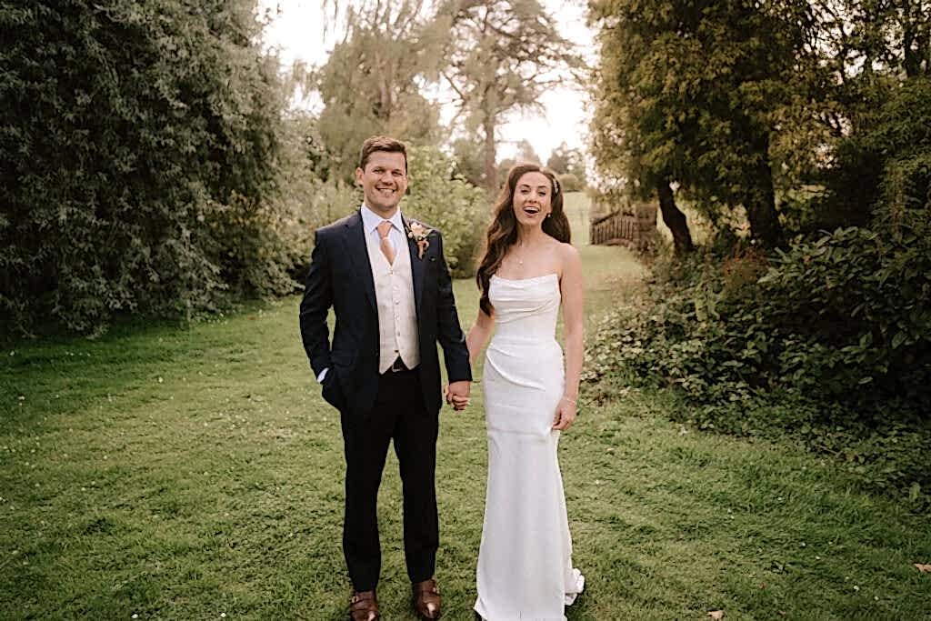 A bride and groom, captured in exquisite wedding photography, stand together on a grassy path surrounded by trees. The groom wears a navy suit, and the bride is radiant in her white dress. Smiling and holding hands, they are framed by a lush, green backdrop. Image by Pearce Wedding Photography.