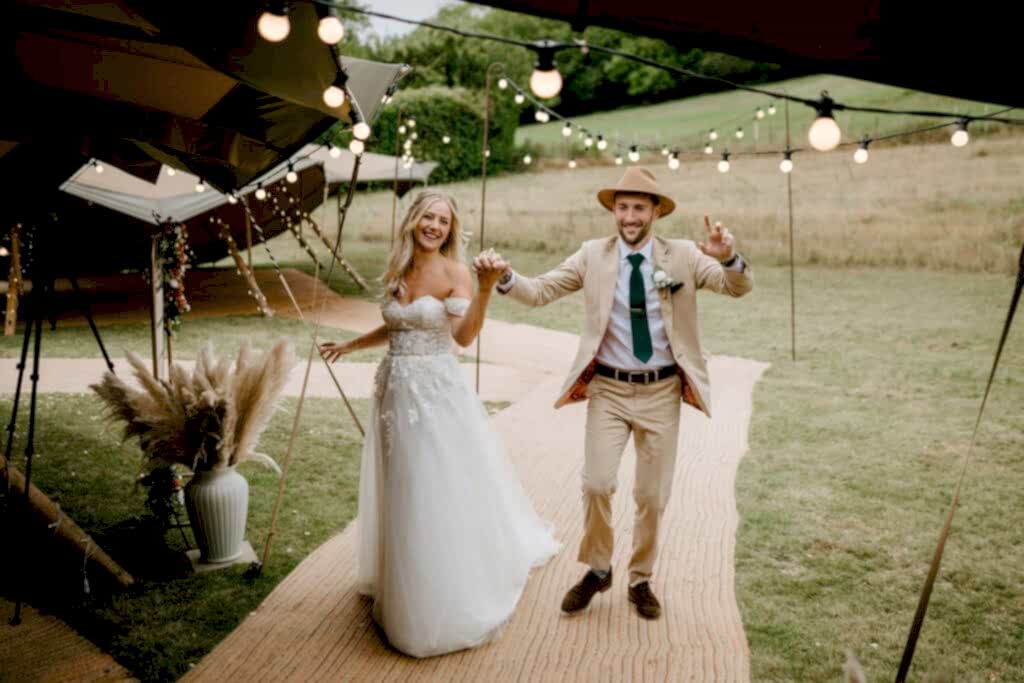 A bride and groom joyfully walk hand in hand under festive string lights, beautifully captured by a wedding photographer in Kent. The bride wears a white gown, and the groom dons a beige suit with a hat. They are outdoors, surrounded by greenery and a decorated tent. Image by Pearce Wedding Photography.