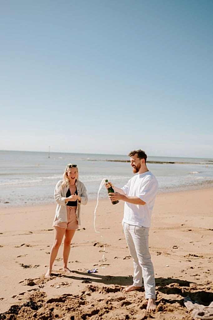 A man and a woman are standing on a sandy beach at Viking Bay. The man, holding a champagne bottle, is popping it open, releasing a spray of champagne. The woman stands nearby, smiling and watching the celebration. The ocean is visible in the background under a clear sky, perfect for their wedding photographer to capture the moment. Image by broadstairs wedding photographer pearce wedding photography