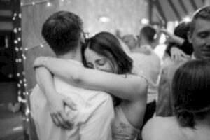 A black and white photo captured by a Kent wedding photographer shows a couple embracing tightly in a warm hug on the dance floor. The setting is festive, with blurred lights and joyful guests dancing in the background. The woman wears glasses and smiles softly, radiating happiness and affection. Image by Pearce Wedding Photography.