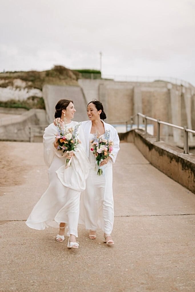 Two women in white dresses holding flowers, smiling.