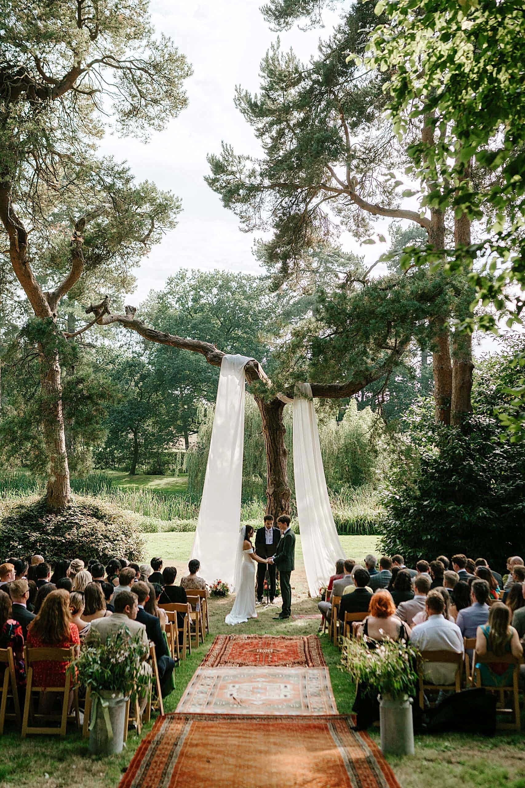 A couple stands at a rustic outdoor wedding altar framed by trees and flowing white fabric, captured perfectly by a Kent wedding photographer. Guests sit on wooden chairs, lining both sides of an aisle adorned with rugs. The ceremony unfolds in a scenic, forested setting under a serene sky. Image by Pearce Wedding Photography.