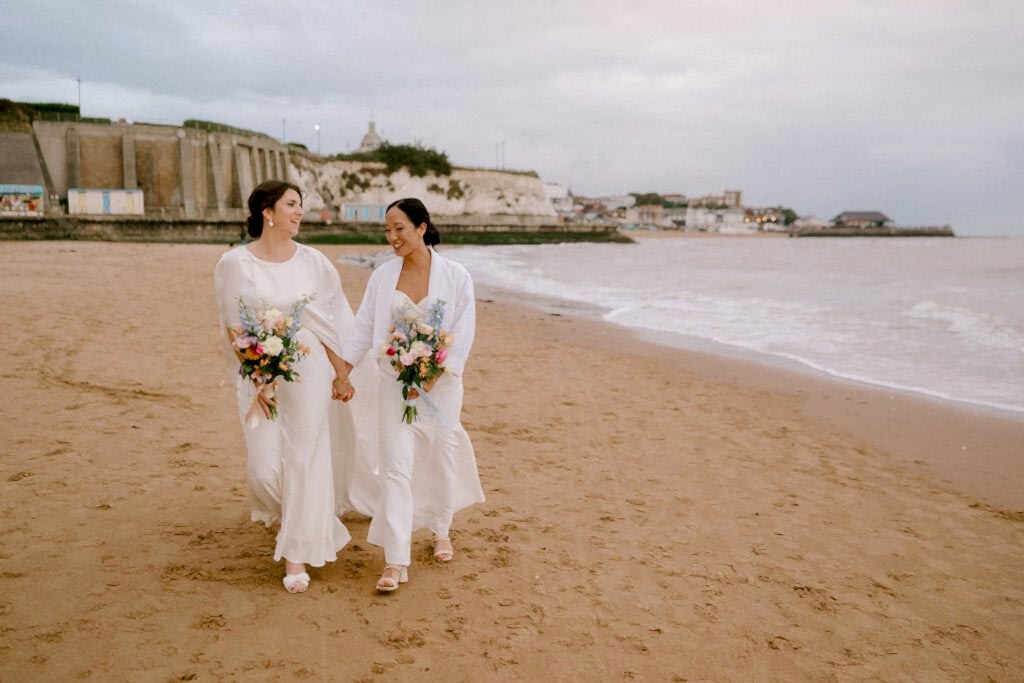 Two people in white outfits walk hand in hand on a sandy Broadstairs beach, each holding a vibrant bouquet of flowers. The backdrop features coastal buildings and an overcast sky, enhancing the serene and intimate wedding atmosphere. Image by Pearce Wedding Photography.